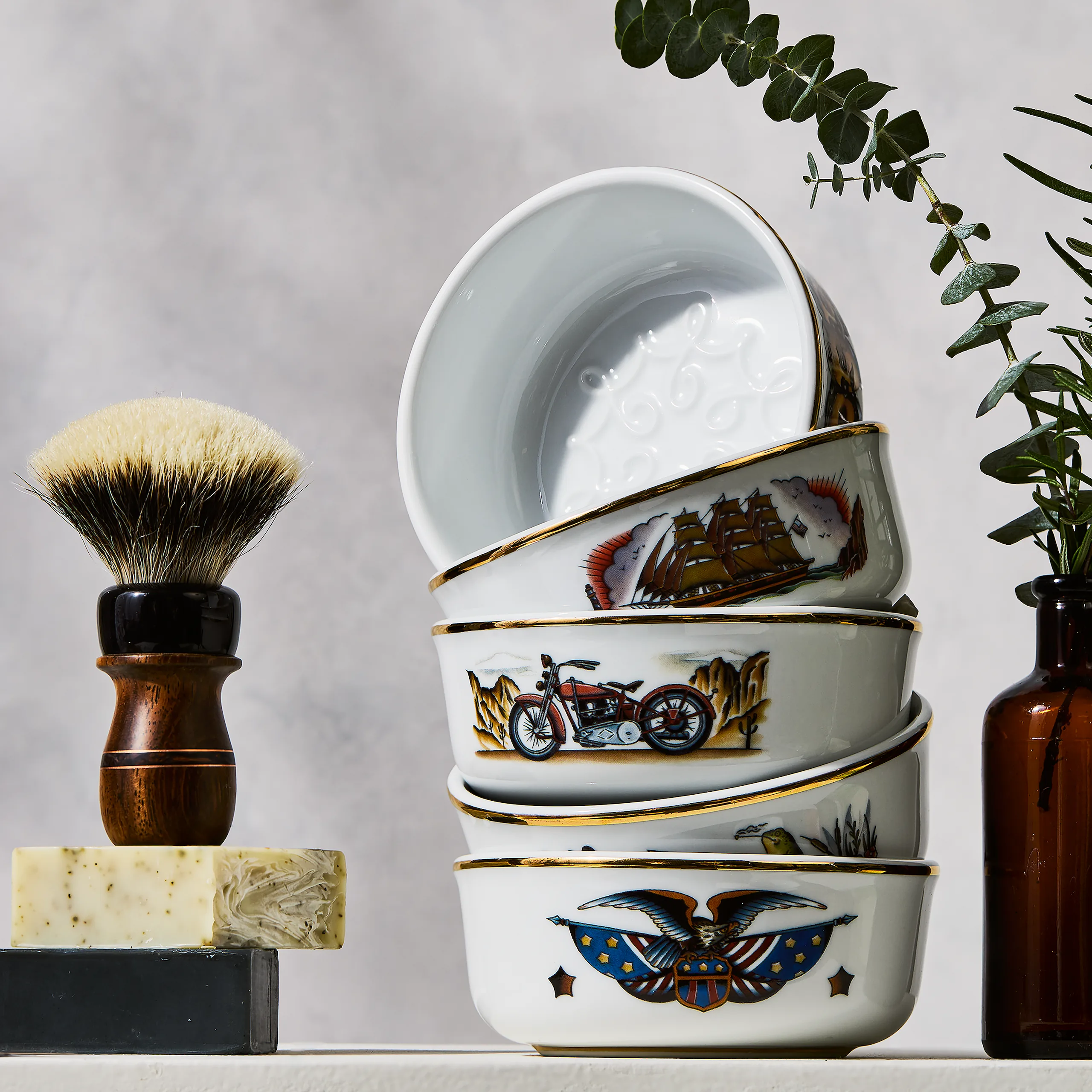 Stacked porcelain shaving bowls featuring original illustrations by Laurent Trelaun, alongside shaving brush and foliage