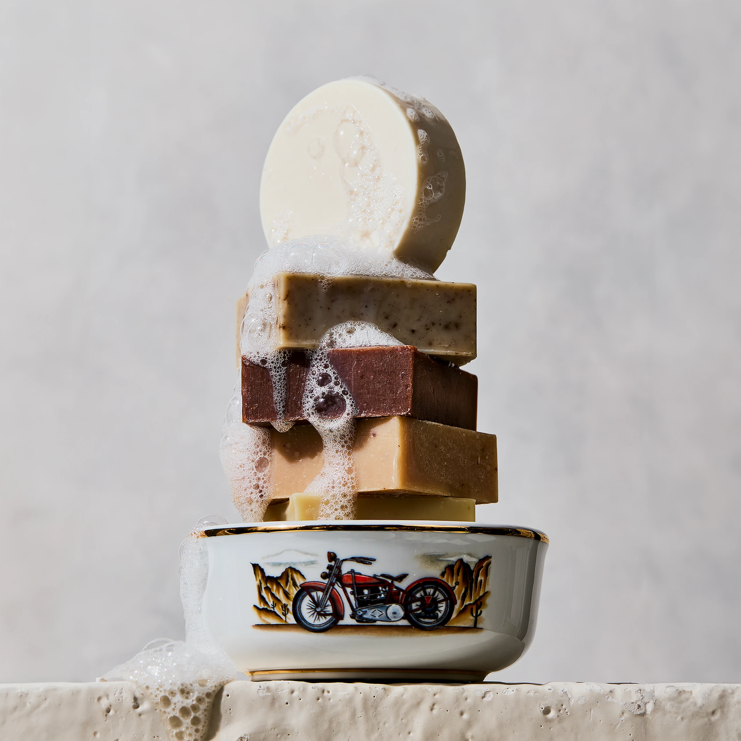 Porcelain shaving bowl with vintage Harley-Davidson illustration stacked with handmade soap bars and shaving lather, set against a light grey patterned backdrop