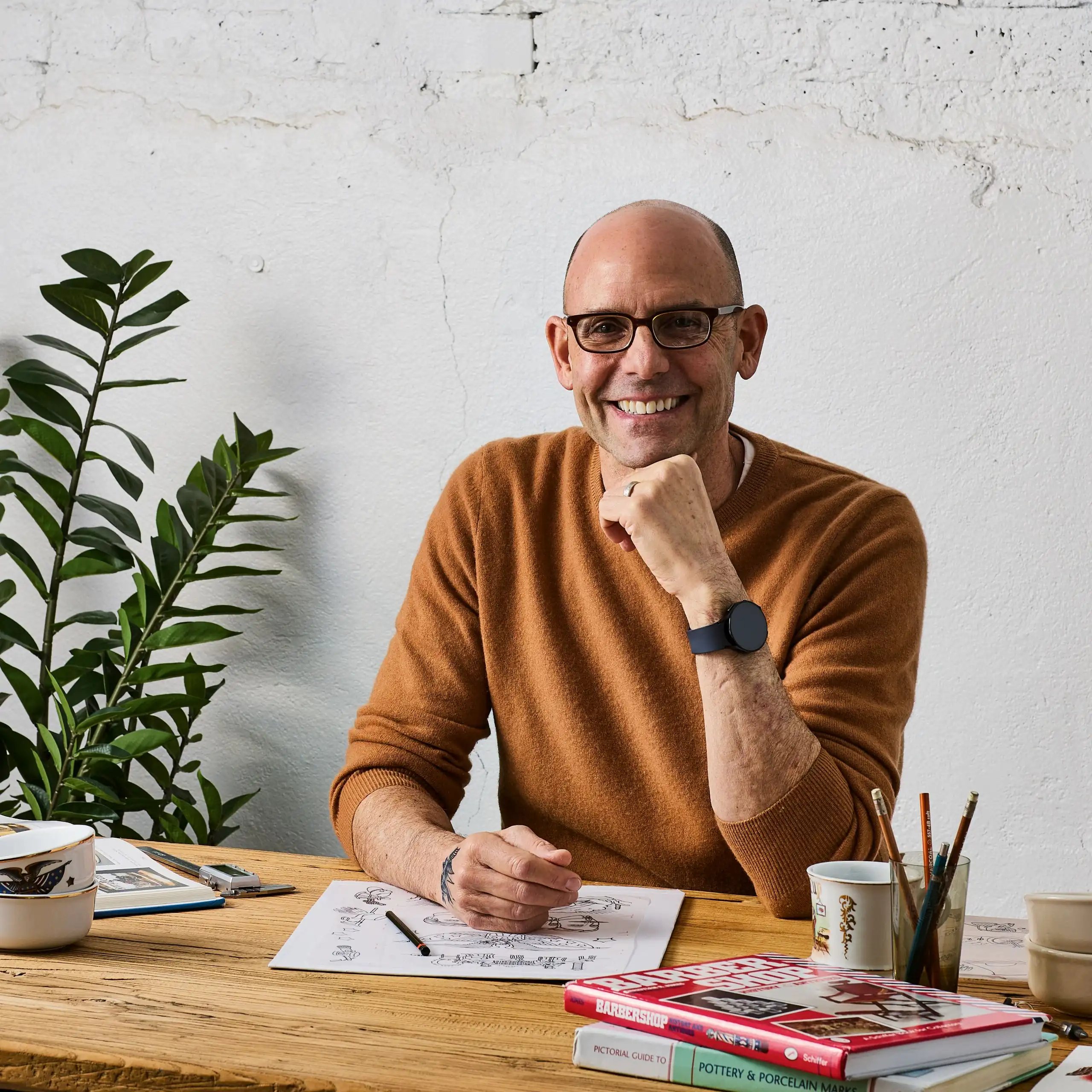 David Altshuler, founder of Brousseau & Dov, seated at a design table with sketchbooks and ceramic references, smiling in a ginger-colored sweater
