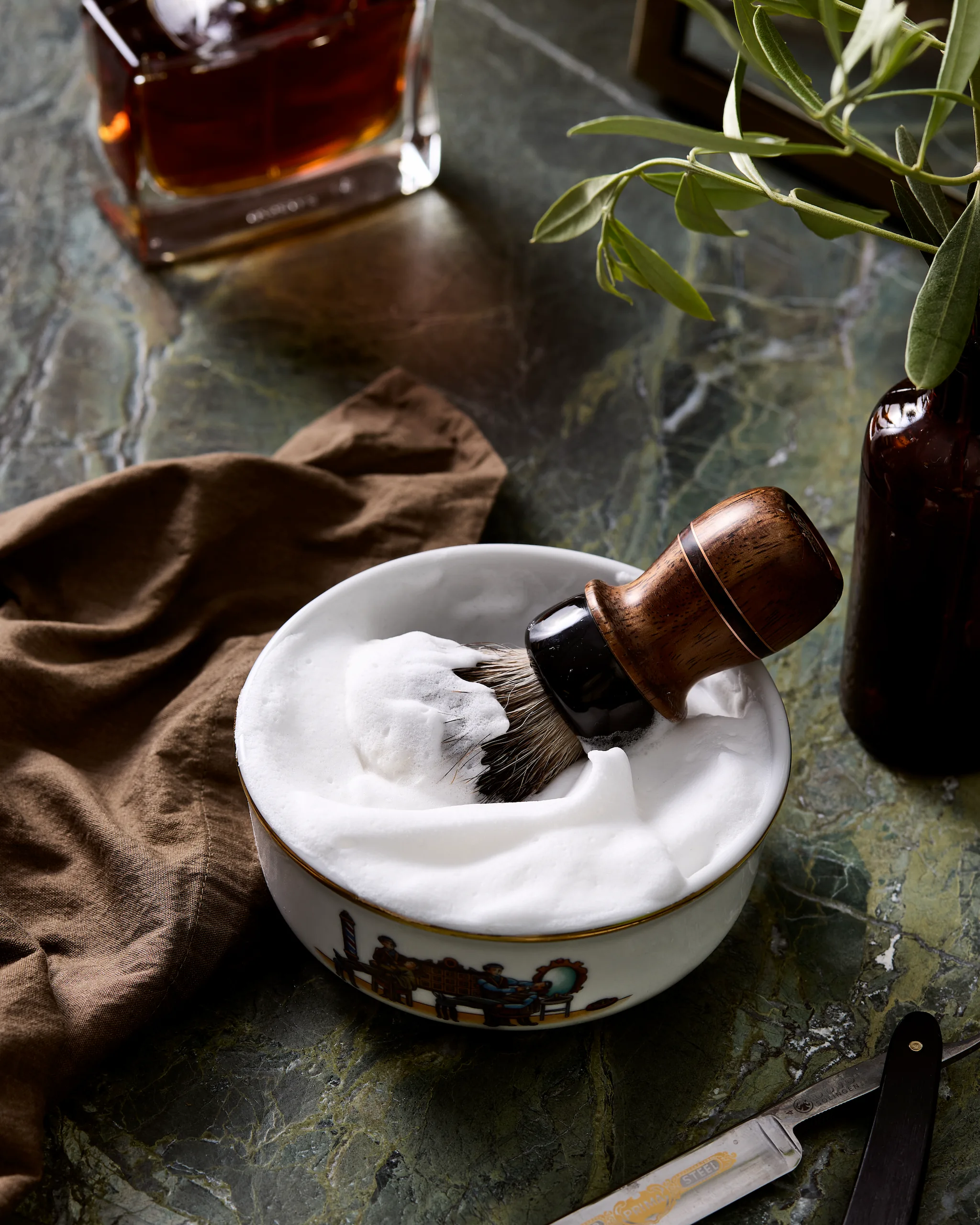 Shaving brush resting in fully developed lather inside porcelain shaving bowl on marble surface, with brown towel, straight razor, and perfume bottle nearby