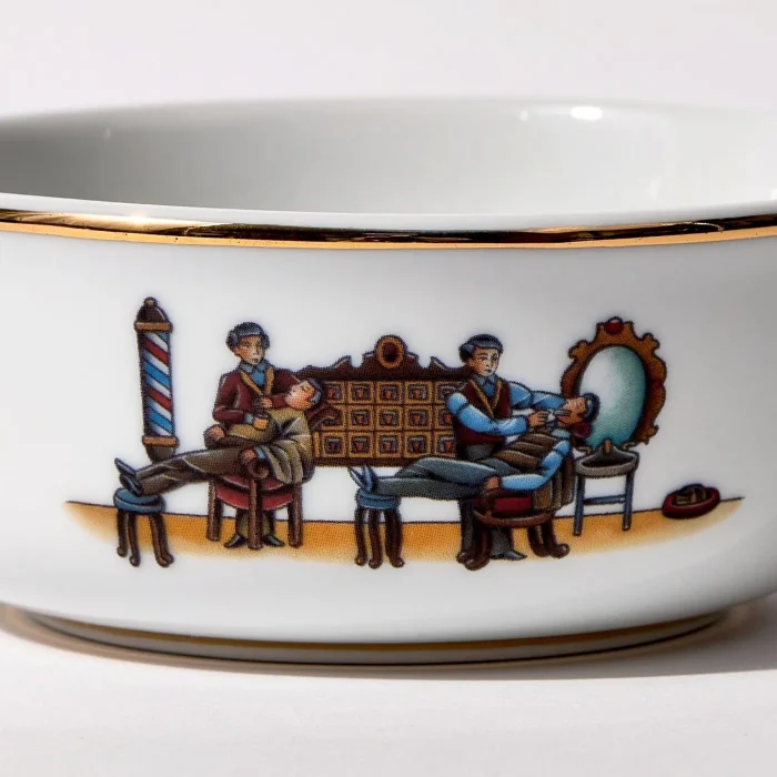 Detail view of porcelain shaving bowl with illustration of vintage barbershop scene, gold trim