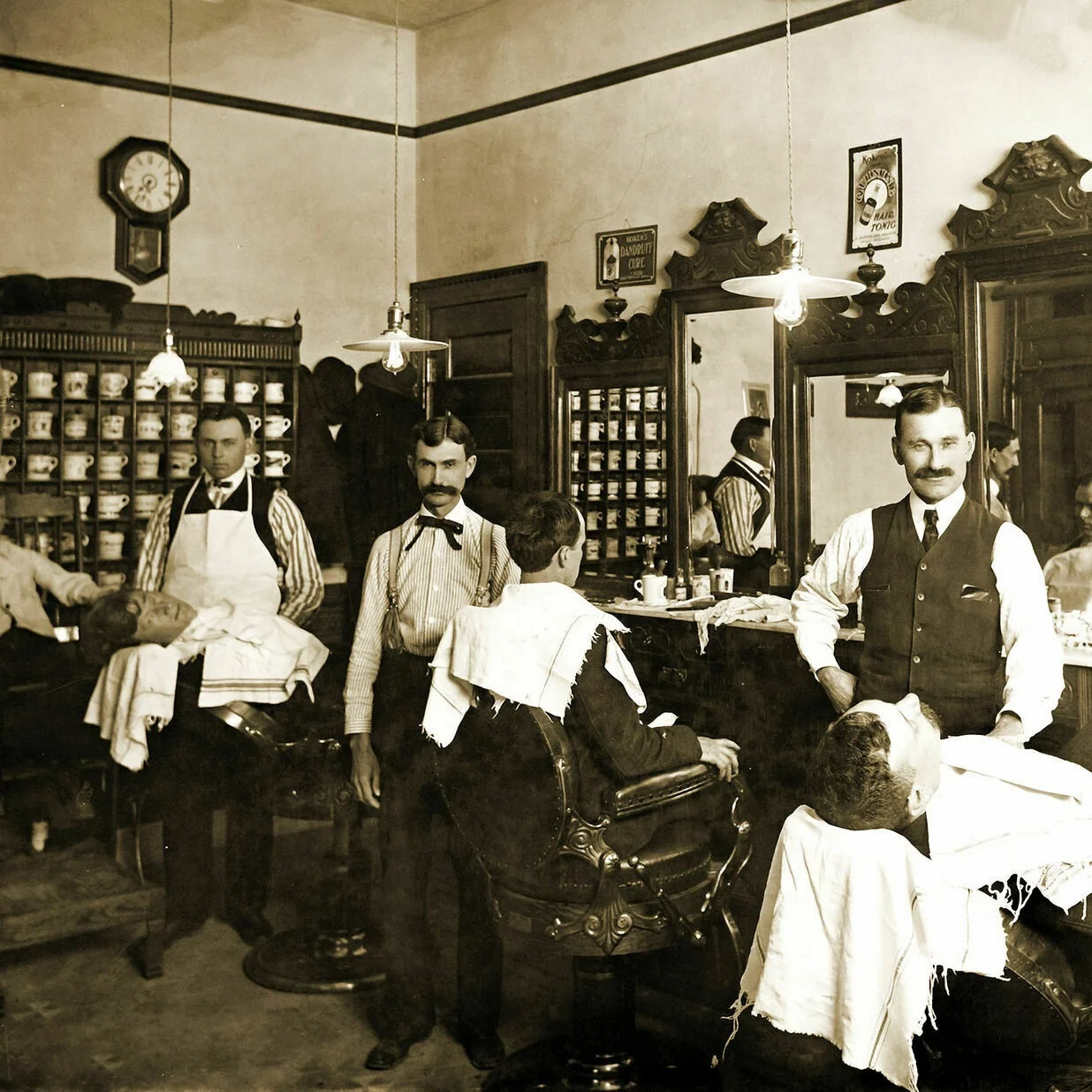 Sepia photograph of early 20th-century barbershop with barbers shaving clients and shelves filled with personalized porcelain shaving mugs