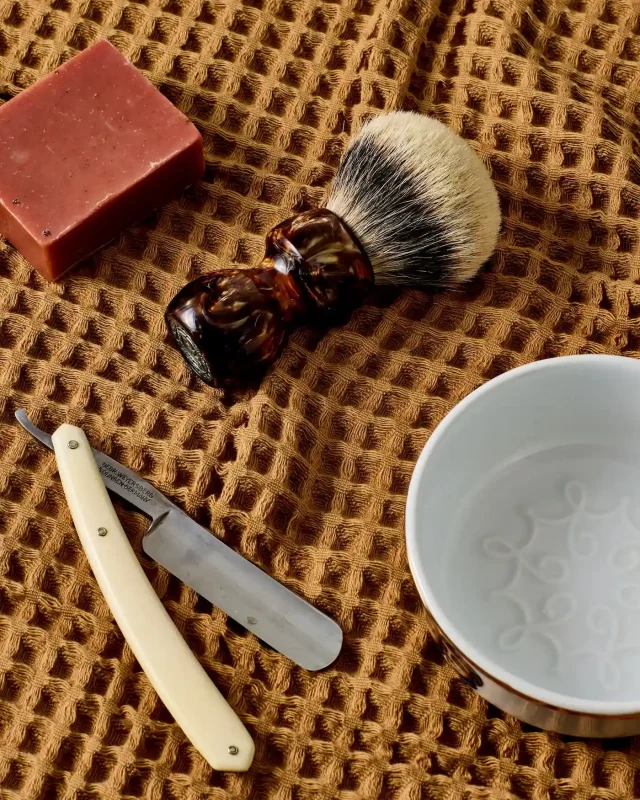 Shaving brush, straight razor, and porcelain bowl arranged on textured towel alongside a bar of soap, illustrating traditional shaving care