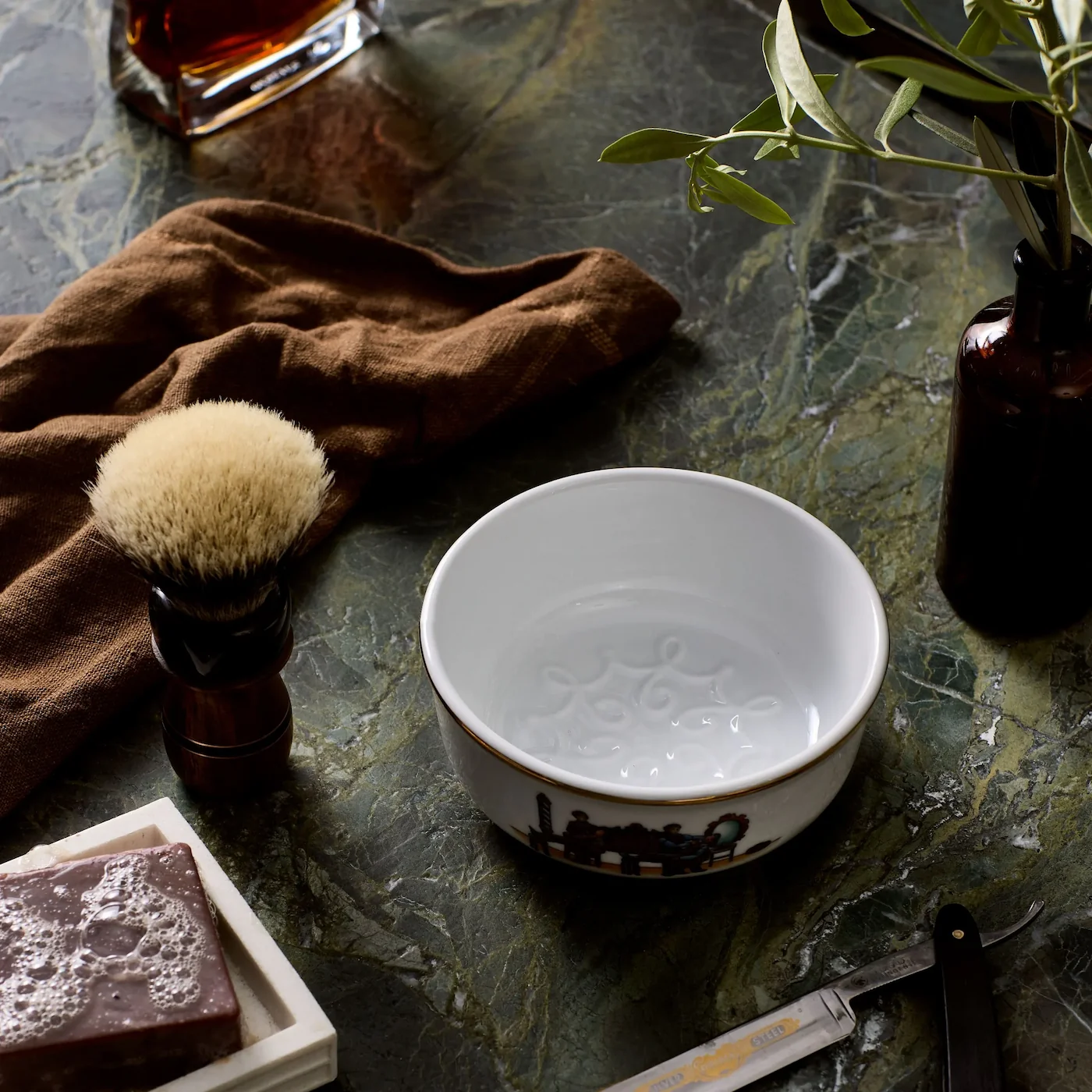 Porcelain shaving bowl on marble surface, with brown towel, shaving brush, soap, straight razor, and perfume bottle nearby