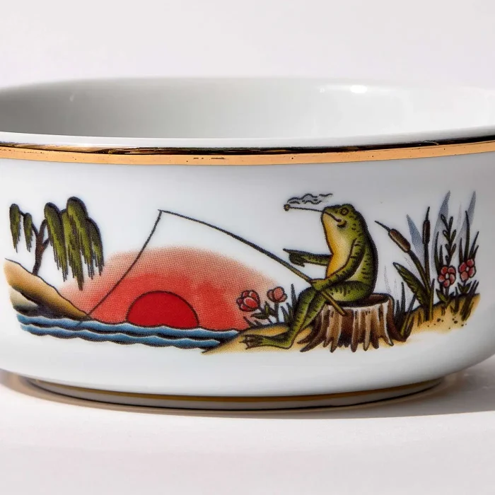 Detail view of porcelain shaving bowl with illustration of anthropomorphic frog fishing and riverside backdrop, gold trim