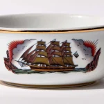 Detail view of porcelain shaving bowl with illustration of vintage sailing ship and port backdrop, gold trim