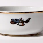 Detail view of back of shaving bowl with illustration of vintage captain's hat, sextant, and coiled rope, gold trim