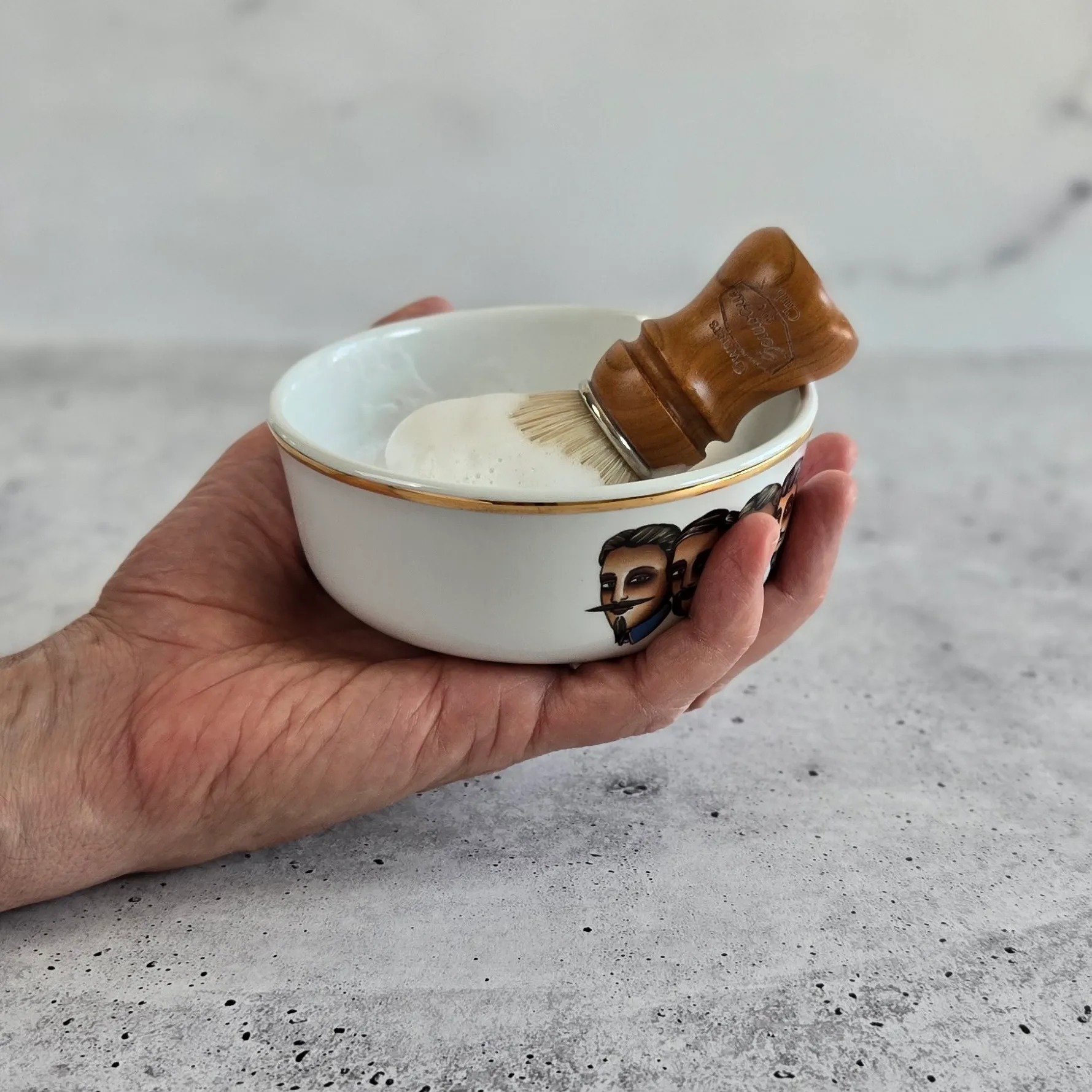 Hand holding a porcelain shaving bowl with shaving brush resting inside